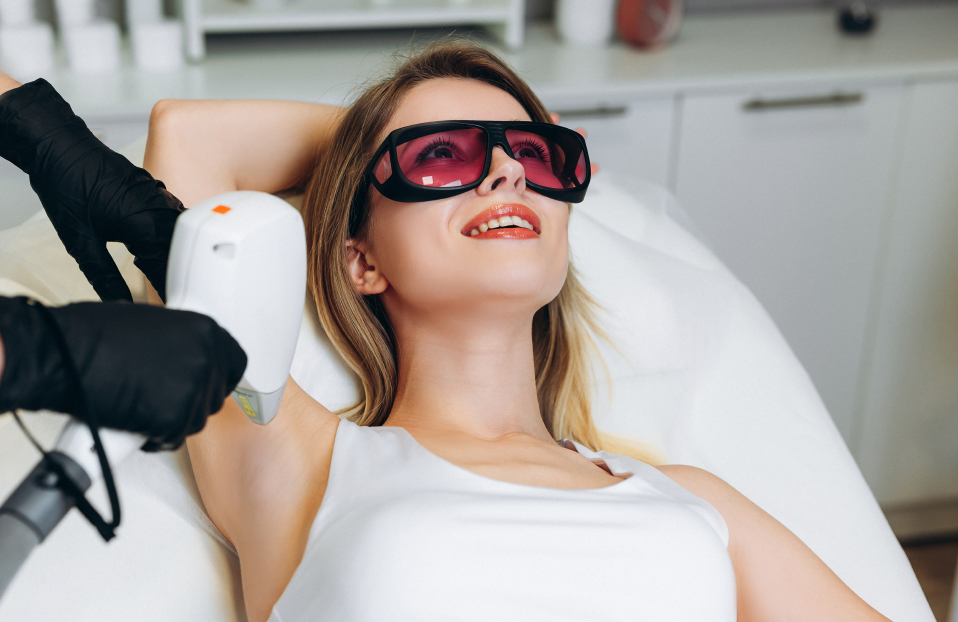 Female cosmetician in uniform using machine for laser hair removal in room with client lying on medical table in beauty clinic